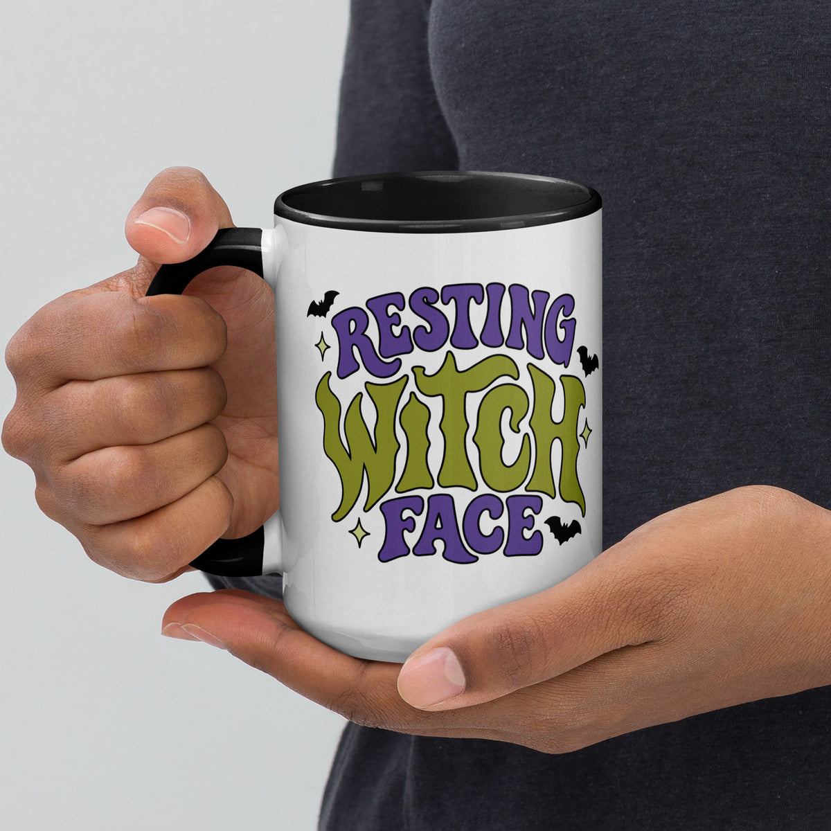 A person holding a white ceramic mug with a black handle and interior. The mug features a colorful graphic that reads 'RESTING WITCH FACE' in purple and green lettering with small black bat silhouettes, against a light gray background and dark gray shirt.