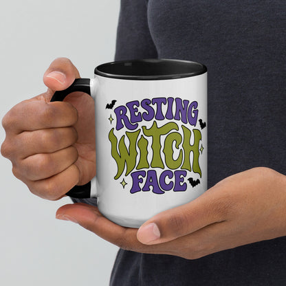 A person holding a white ceramic mug with a black handle and interior. The mug features a colorful graphic that reads 'RESTING WITCH FACE' in purple and green lettering with small black bat silhouettes, against a light gray background and dark gray shirt.