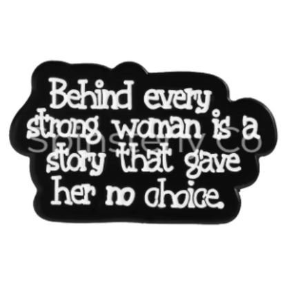 A black enamel pin with white text that reads, 'Behind every strong woman is a story that gave her no choice.' on a plain white background.