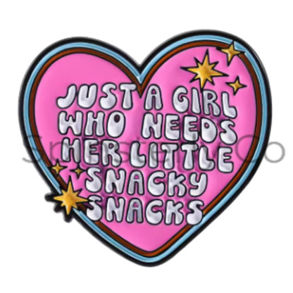 A heart-shaped enamel pin with a pink background, featuring the text 'JUST A GIRL WHO NEEDS HER LITTLE SNACKY SNACKS' in white bubble letters with black outlines. The pin is decorated with gold starbursts and has a blue and brown border, set against a plain white background.