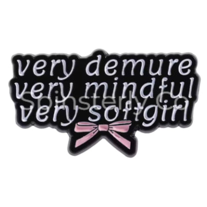 A black enamel pin with white cursive text reading 'very demure very mindful very softgirl' and a pink bow at the bottom, shown on a plain white background.