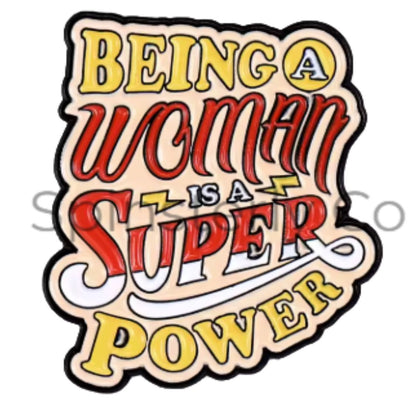 A die-cut lapel pin with the phrase 'BEING A WOMAN IS A SUPER POWER' in stylized lettering. The design has a cream background with red, yellow, and white text outlined in black, plus small lightning bolt accents.
