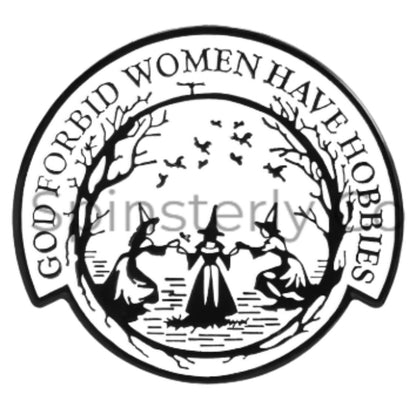 A circular black and white graphic enamel lapel pin featuring two witches in pointed hats standing in a field with bare trees, birds, and a cross above. The outer banner reads 'GOD FORBID WOMEN HAVE HOBBIES' in raised lettering.