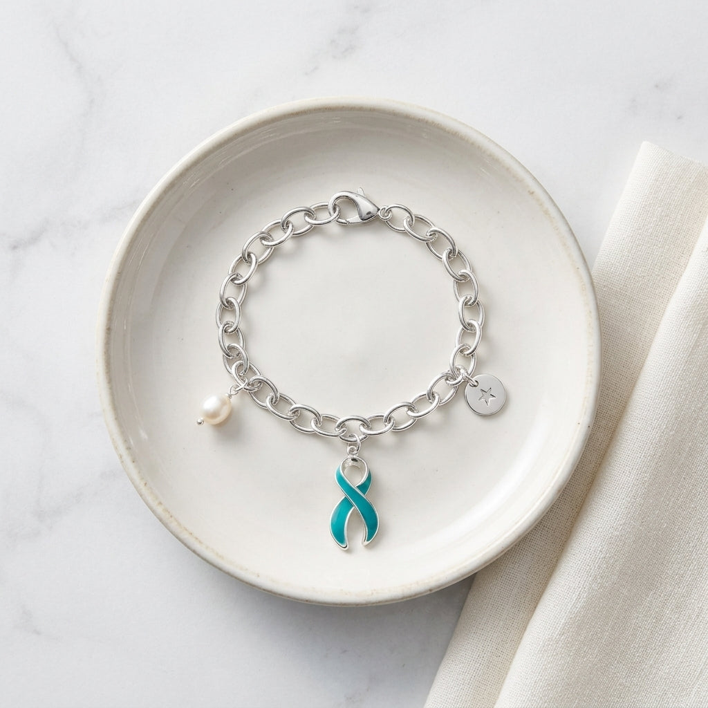 A silver chain bracelet with a teal awareness ribbon charm, a small white pearl bead, and a silver star charm, displayed on a round off-white ceramic dish over a light marble surface with a cream fabric napkin.