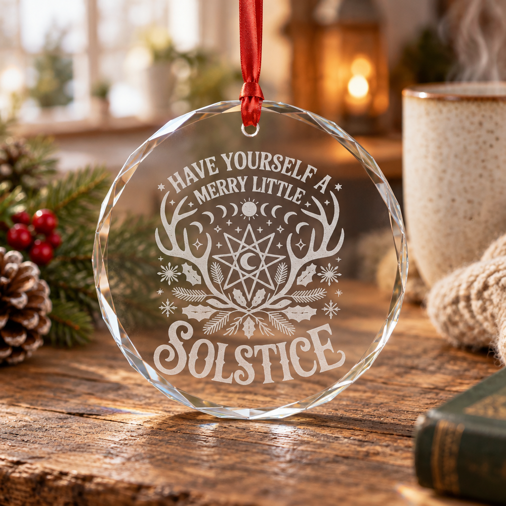 A clear faceted glass or acrylic solstice ornament hanging from a red ribbon, printed with a celestial antler-and-moon design and the words “HAVE YOURSELF A MERRY LITTLE SOLSTICE,” displayed on a rustic wooden table with holiday greenery, a mug, and a candle in the background.