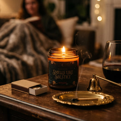A lit candle in a glass jar sits on a wooden table in a cozy living room, with a blurred person reading on the couch in the background. The candle jar has a label that reads 'SMELLS LIKE SOLSTICE'.