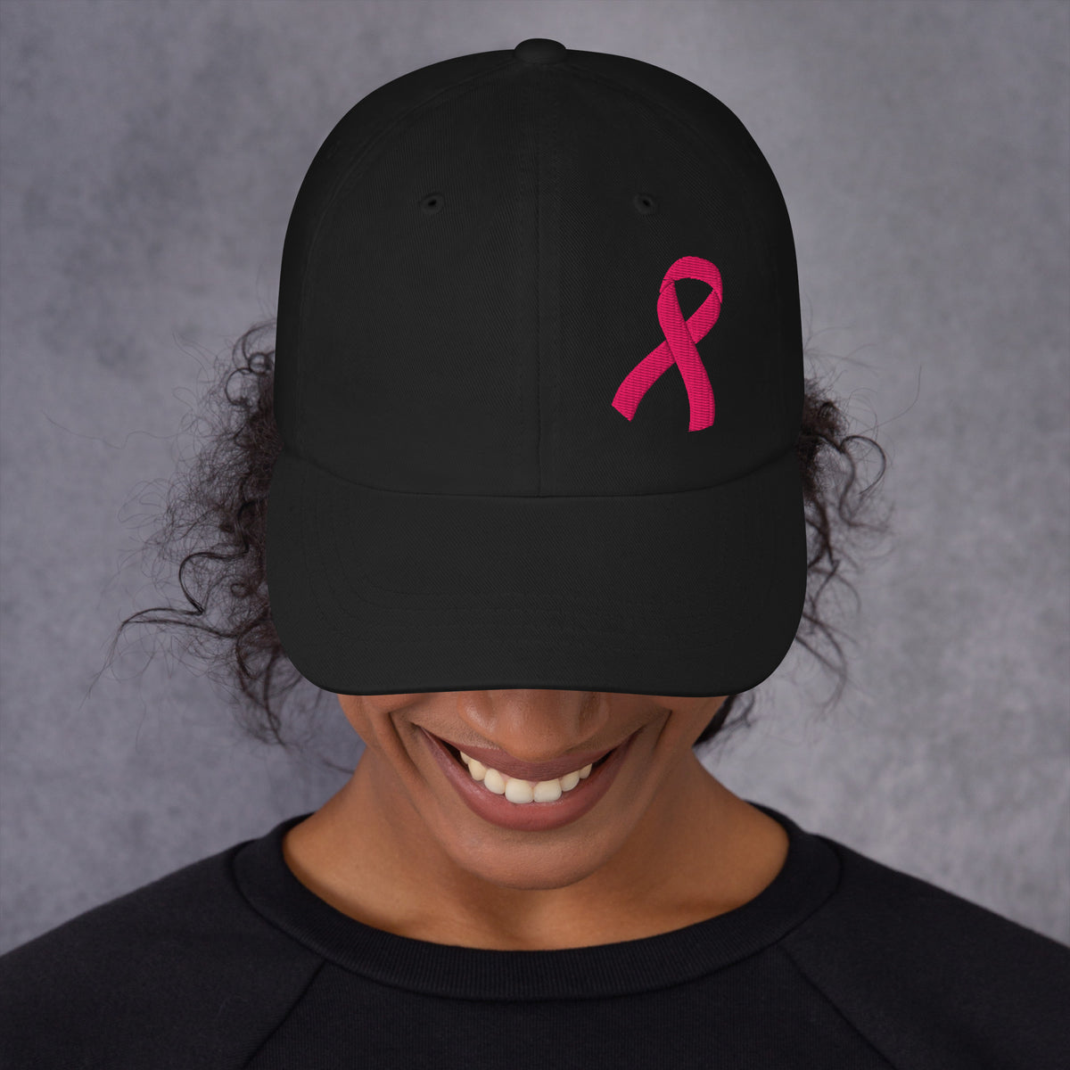 A person wearing a black baseball cap with a embroidered pink ribbon embroidered on the side, shown from the chest up against a textured gray background.