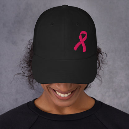 A person wearing a black baseball cap with a embroidered pink ribbon embroidered on the side, shown from the chest up against a textured gray background.