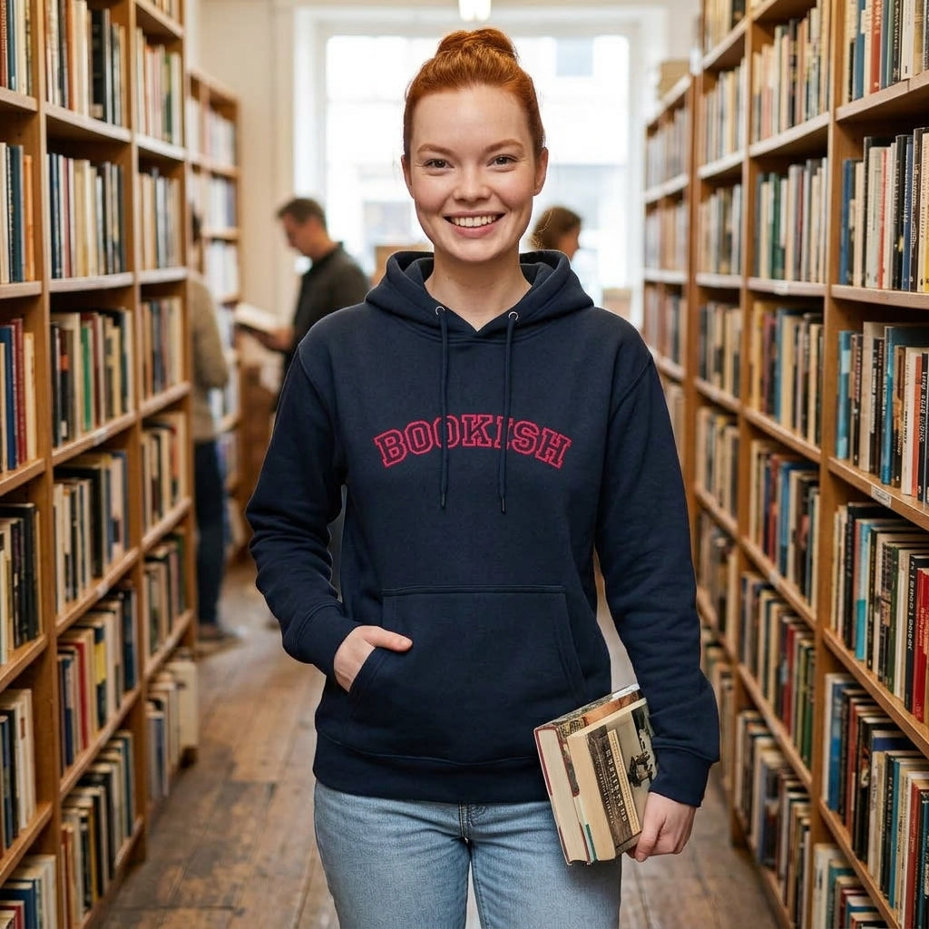A smiling young woman stands in a library aisle wearing a navy hoodie with 'BOOKISH' printed in red across the chest, paired with light blue jeans and holding a book.