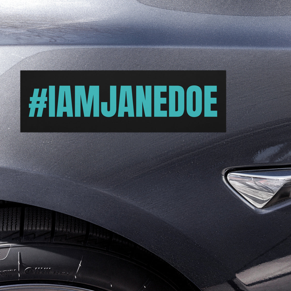A rectangular black bumper sticker with the text '#IAMJANEDOE' in bright teal lettering, shown applied to the side fender of a dark gray car.