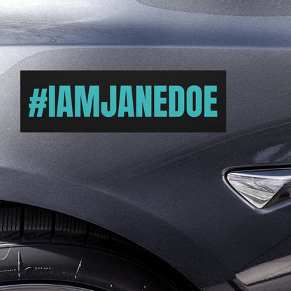 A rectangular black bumper sticker with the text '#IAMJANEDOE' in bright teal lettering, shown applied to the side fender of a dark gray car.