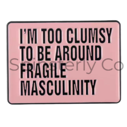 A rectangular pink enamel pin with a black border and bold black text reading 'I'M TOO CLUMSY TO BE AROUND FRAGILE MASCULINITY'. 