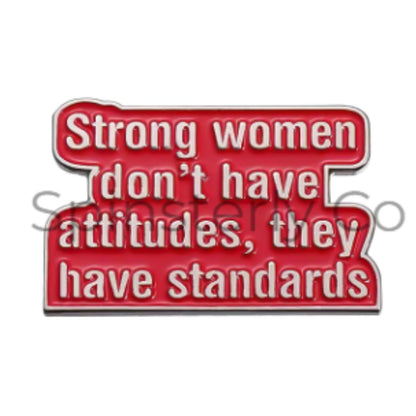 A red enamel pin with white text that reads 'Strong women don't have attitudes, they have standards' on a plain white background.