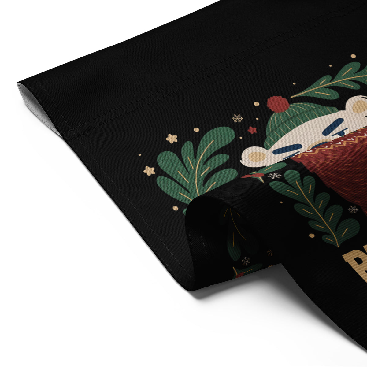 A close-up of a black fabric product, likely a blanket or towel, featuring a festive holiday illustration of a sleeping bear in a green knit hat surrounded by green leaves and gold stars.