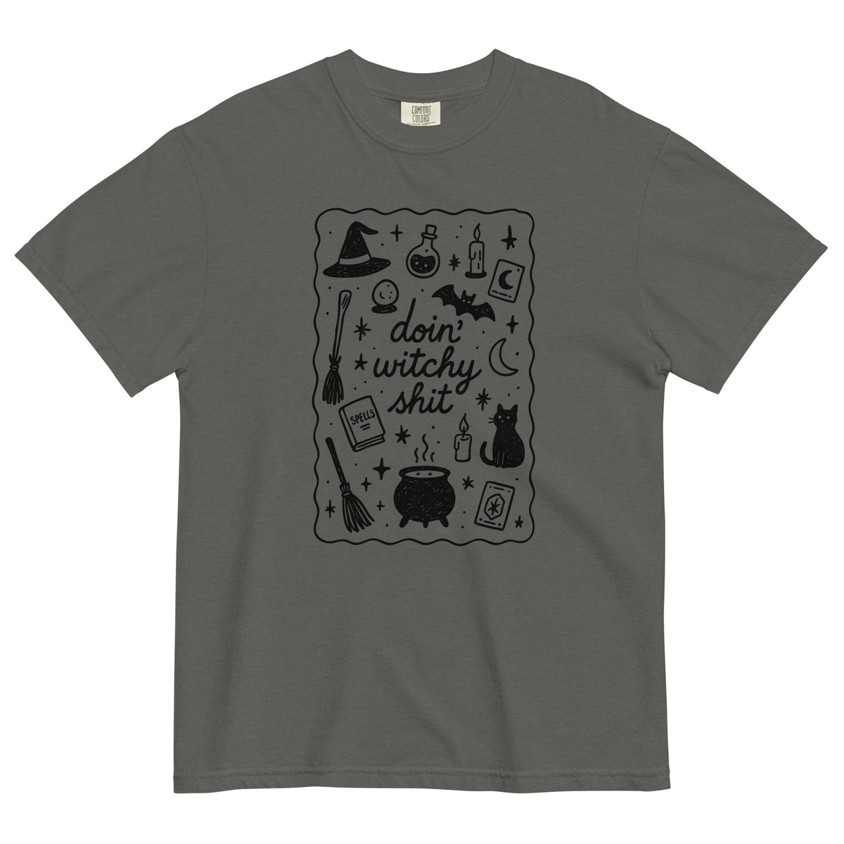 A grey, heavyweight cotton t-shirt, with a black scatter graphic that says 'doin' witchy shit' with Halloween inspired iconography design which includes witch hats, cauldron, black cats, brooms, potions, candles, moon and stars with a squiggle line border around the graphic.