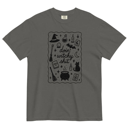 A grey, heavyweight cotton t-shirt, with a black scatter graphic that says 'doin' witchy shit' with Halloween inspired iconography design which includes witch hats, cauldron, black cats, brooms, potions, candles, moon and stars with a squiggle line border around the graphic.