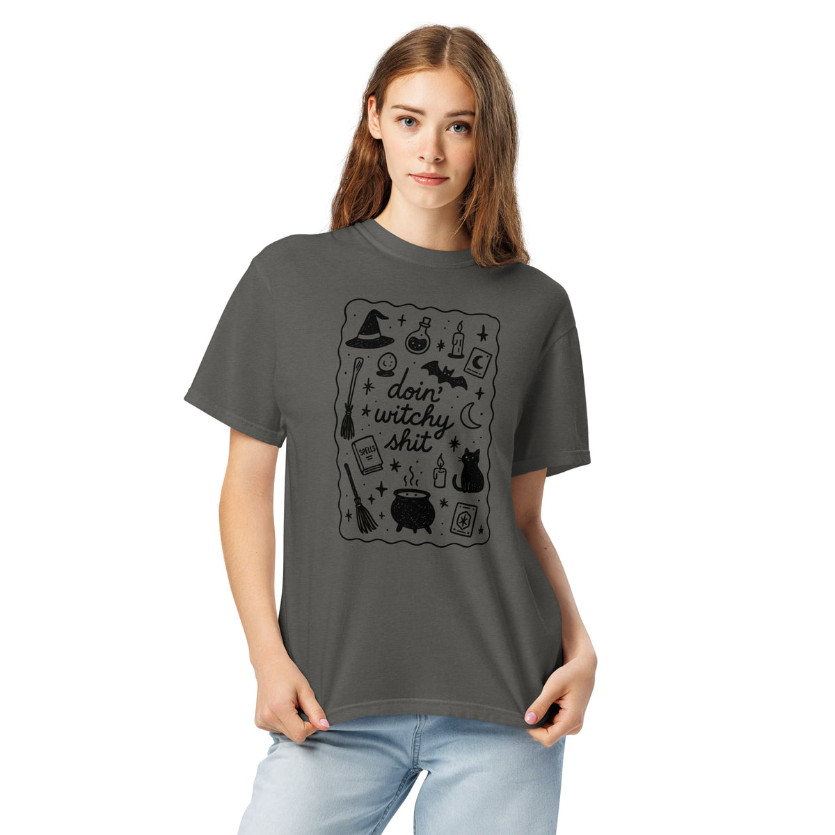 A woman model wearing a heavyweight-cotton t-shirt in grey with a Halloween inspired design that reads 'doin' witchy shit' with scattered Halloween graphics inside a squiggle line border, with a white background.