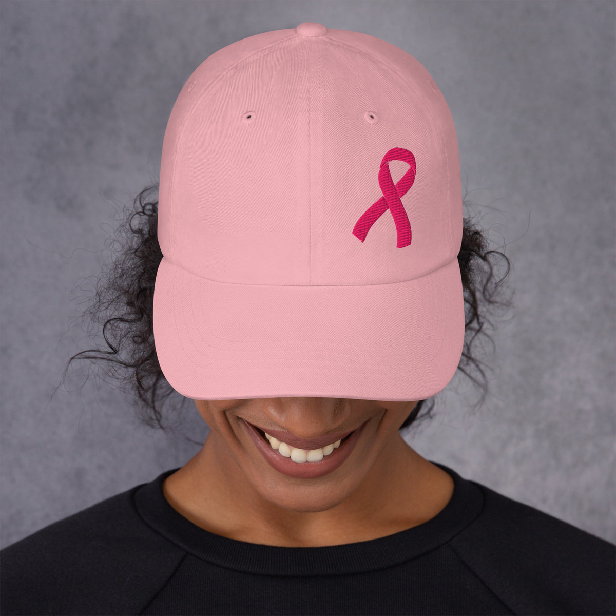 A person wearing a light pink baseball cap with a darker pink embroidered breast cancer awareness ribbon on the front, photographed against a textured gray background.
