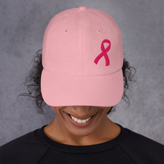 A person wearing a light pink baseball cap with a darker pink embroidered breast cancer awareness ribbon on the front, photographed against a textured gray background.
