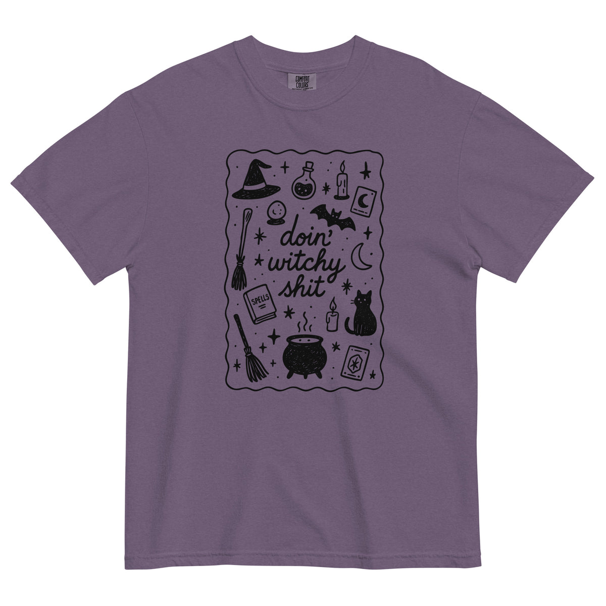 A purple, heavyweight cotton t-shirt, with a black scatter graphic that says 'doin' witchy shit' with Halloween inspired iconography design which includes witch hats, cauldron, black cats, brooms, potions, candles, moon and stars with a squiggle line border around the graphic.