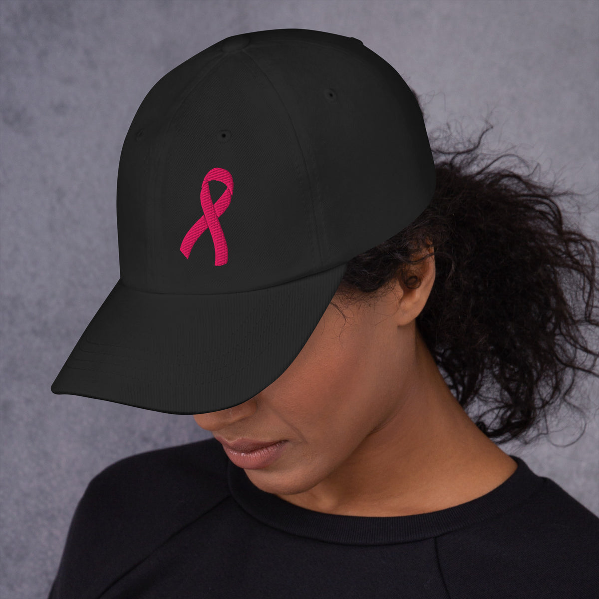 A person wearing a black baseball cap with a pink breast cancer awareness ribbon embroidered on the front, photographed against a textured gray background.