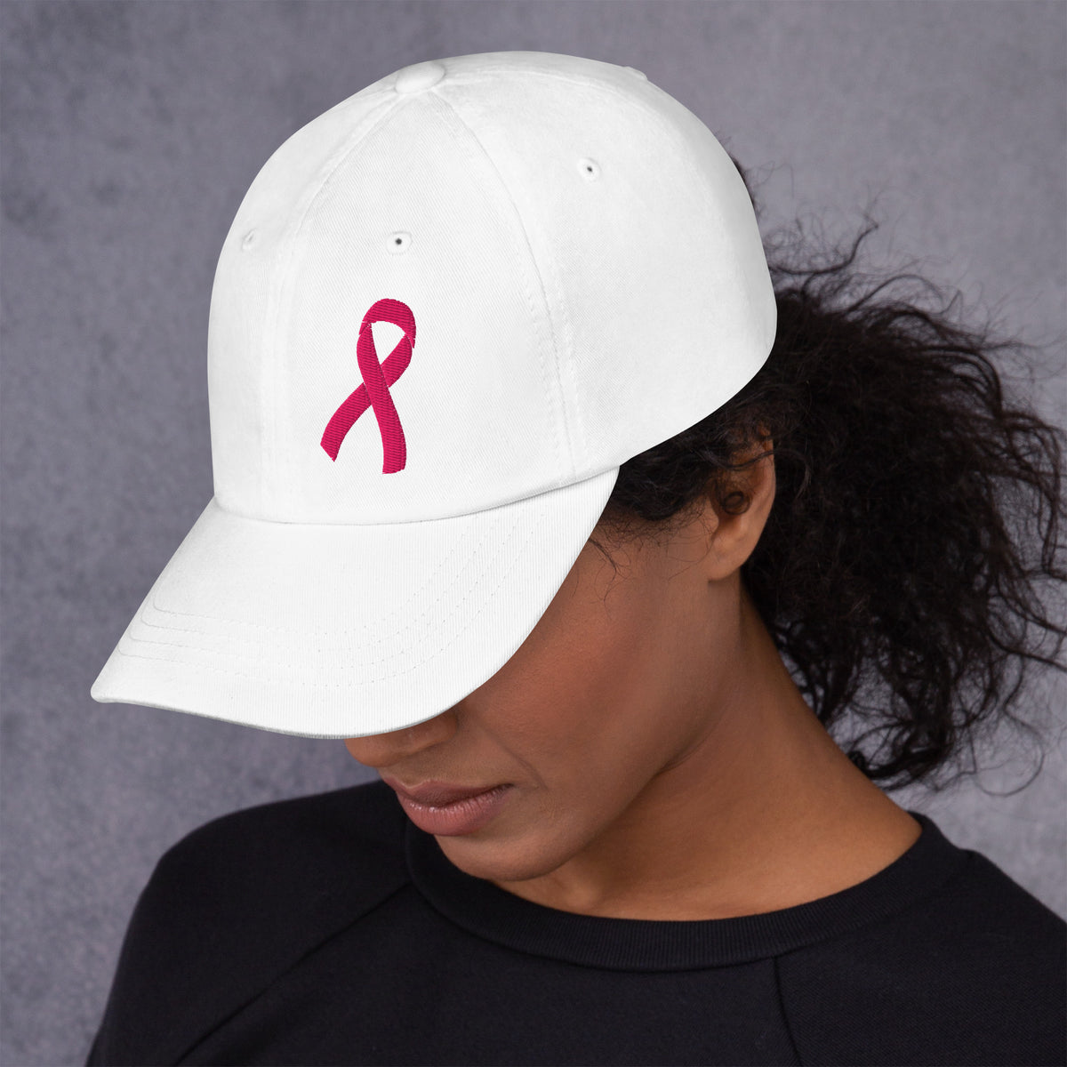 Person wearing a white cap a pink breast cancer awareness ribbon embroidered on the front on a gray background