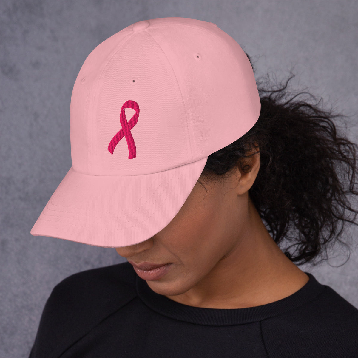 A woman wearing a light pink baseball cap with a dark pink embroidered breast cancer ribbon on the front, shown from the side against a textured gray background.