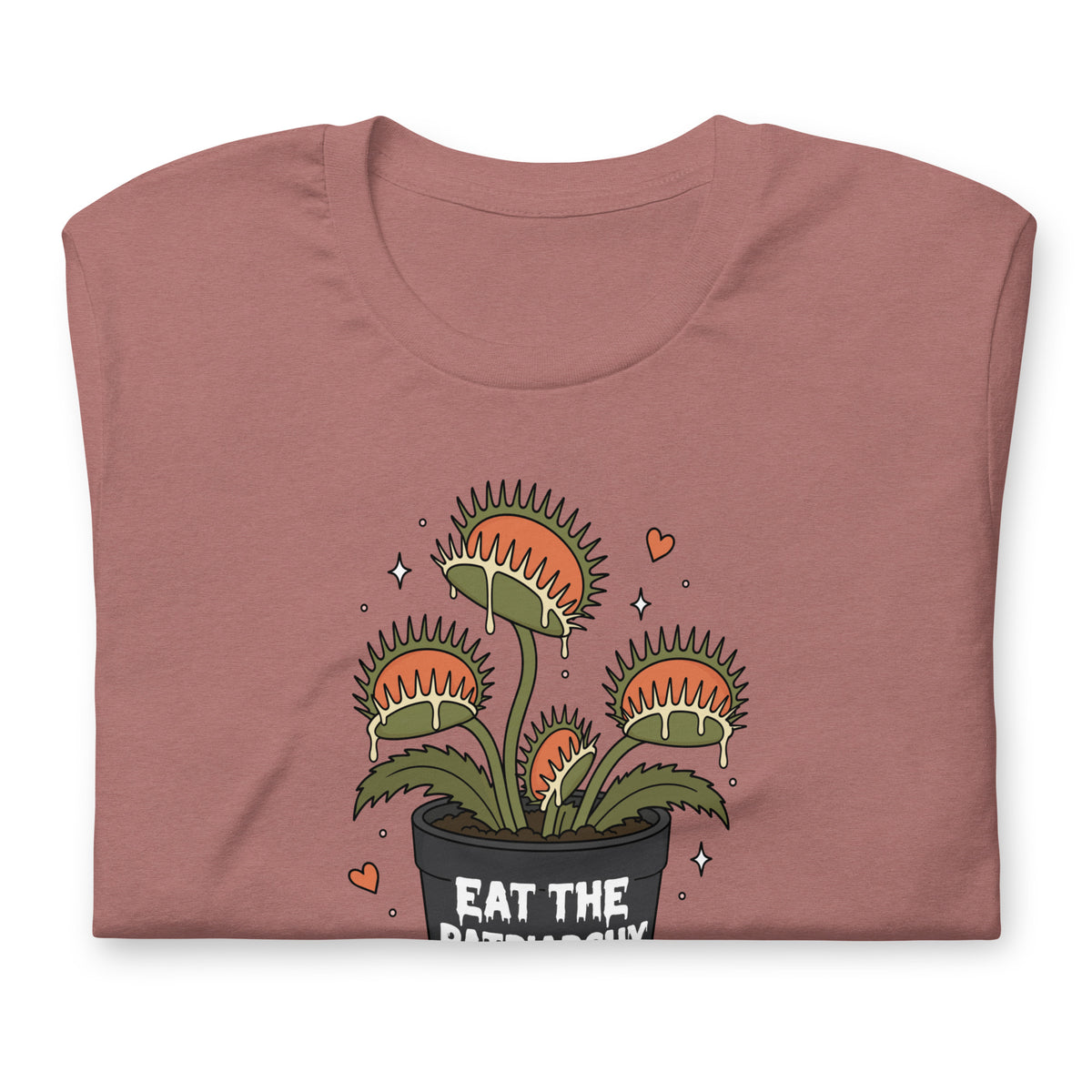 A folded dusty rose t-shirt with a cartoon graphic of a Venus flytrap plant in a black pot with the text 'EAT THE PATRIARCHY' and small orange hearts, shown on a plain white background.