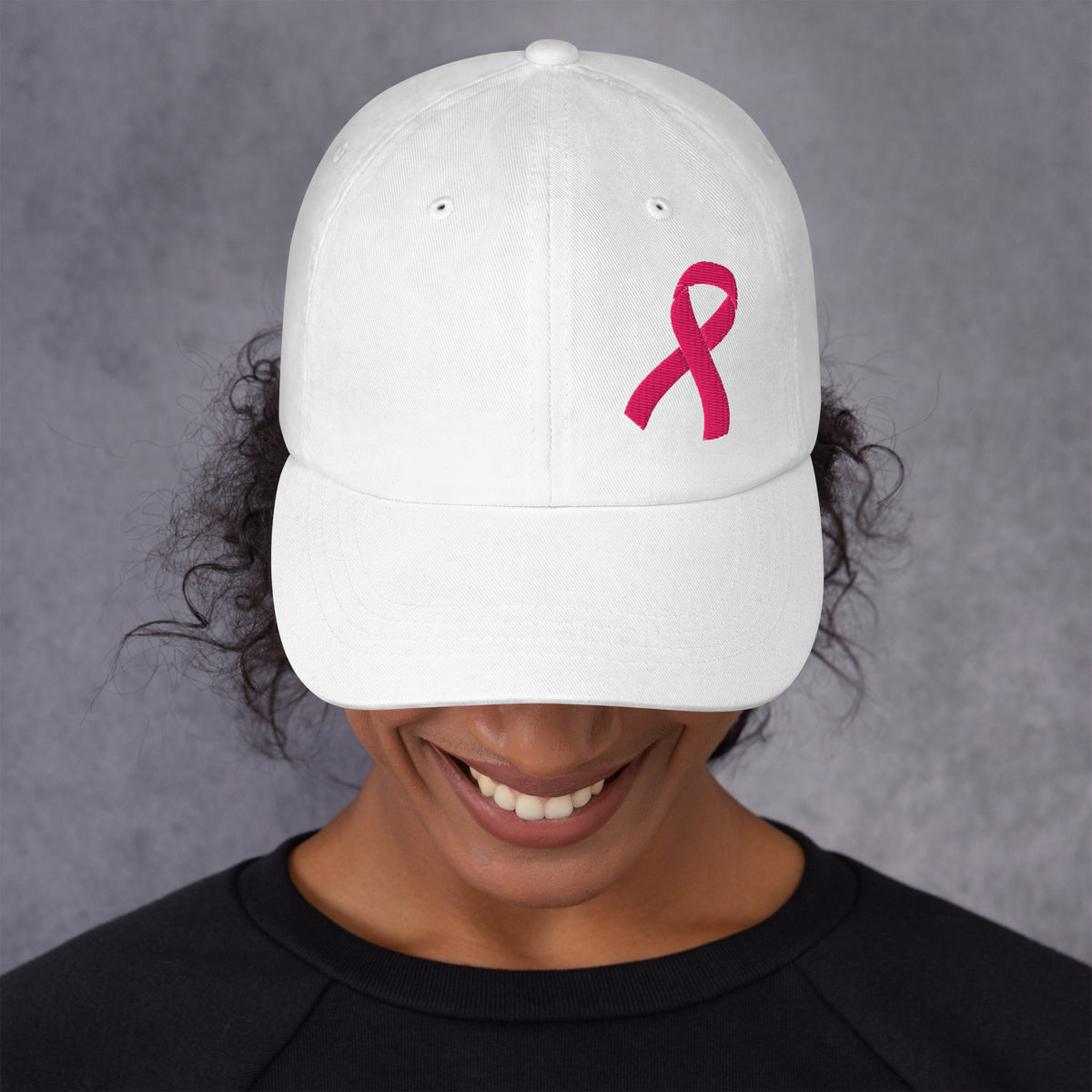 A white baseball cap with a pink breast cancer awareness ribbon embroidered on the front, worn by a smiling person against a textured gray background.